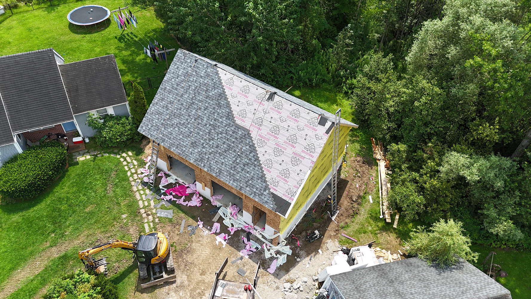 Aerial overhead view of new construction roof installation in Hinesburg Vermont — shingles installed on left plane, ProPink underlayment visible on right, excavator and dump trailer on active job site