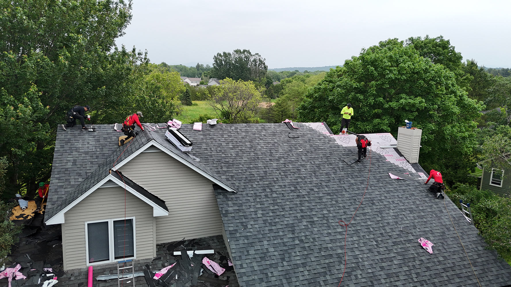 Angled aerial view of Rivers Construction crew installing Owens Corning shingles in Williston Vermont — dormer roofline visible, ProArmor underlayment on upper section, Vermont summer landscape in background