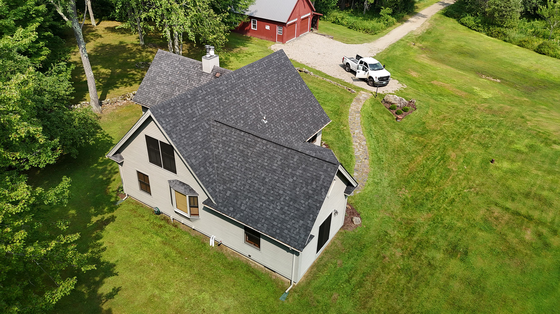 Rear aerial view of completed shingle roof replacement in Stowe Vermont — Rivers Construction truck in driveway, red barn visible, dark charcoal shingles across full roofline